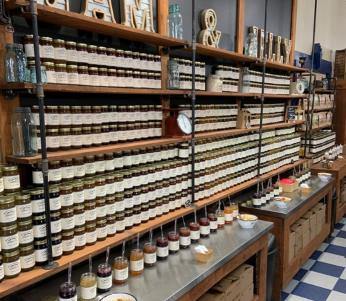 A wooden shelf filled with jars of various jams and preserves, with tasting samples on a table in front.