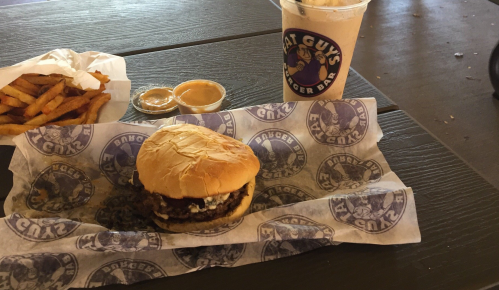 A burger with toppings on a bun, served with fries and a drink, all on a paper-lined table.