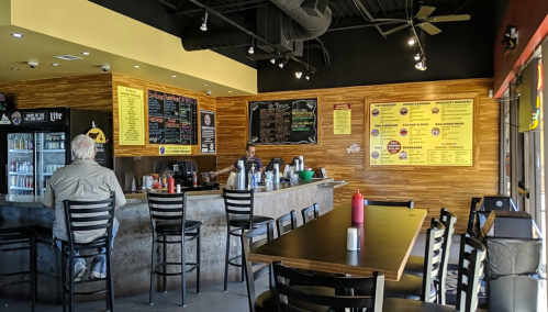 Interior of a casual restaurant with a bar, menu boards, and a few patrons seated at tables and the counter.