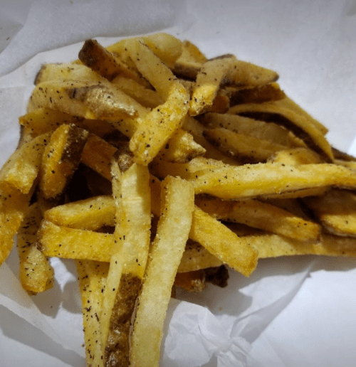 A pile of golden-brown French fries on a white paper towel, lightly seasoned with black pepper.