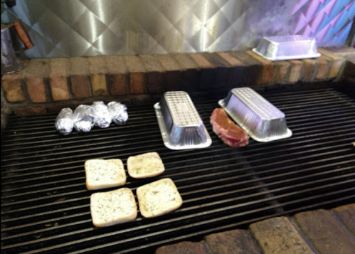 Grill with foil-wrapped potatoes, two metal trays, a piece of meat, and slices of bread on the grates.