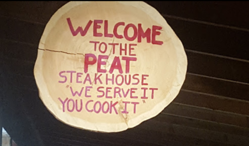 Wooden sign reading "Welcome to the Peat Steak House 'We Serve It, You Cook It'" in bold pink letters.
