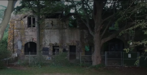 Abandoned stone building partially hidden by trees, surrounded by a fence and overgrown vegetation.