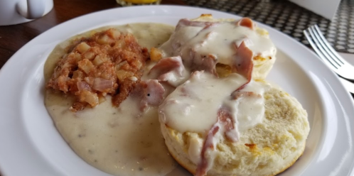 A plate with biscuits and gravy topped with ham, alongside a serving of hash browns in a creamy sauce.