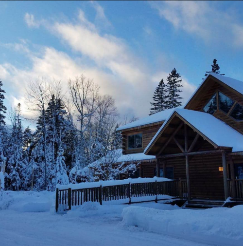 A cozy log cabin surrounded by snow-covered trees under a blue sky with fluffy clouds.