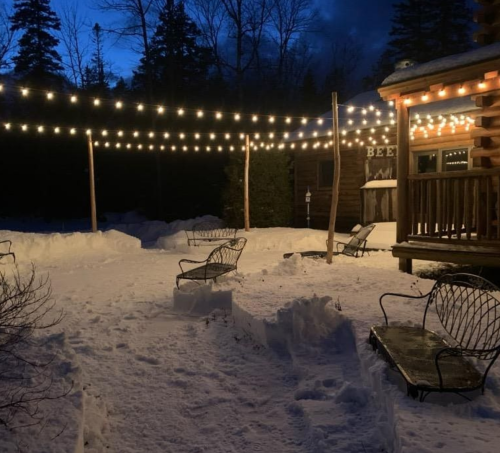 A snowy outdoor area with string lights and benches, illuminated by evening light near a log cabin.
