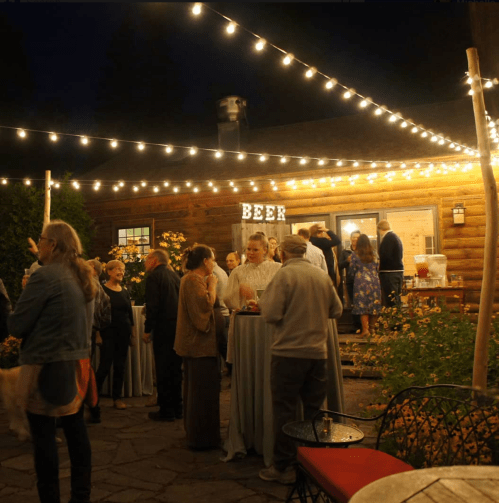 A lively outdoor gathering at night with string lights, people mingling, and a "BEER" sign on a rustic building.