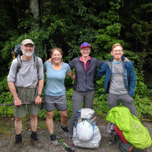 Four hikers stand together in a forested area, smiling and wearing outdoor gear, with backpacks at their feet.