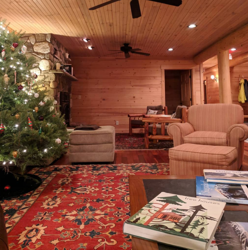 Cozy cabin interior with a Christmas tree, stone fireplace, and books on a colorful rug. Warm wooden walls and furniture.