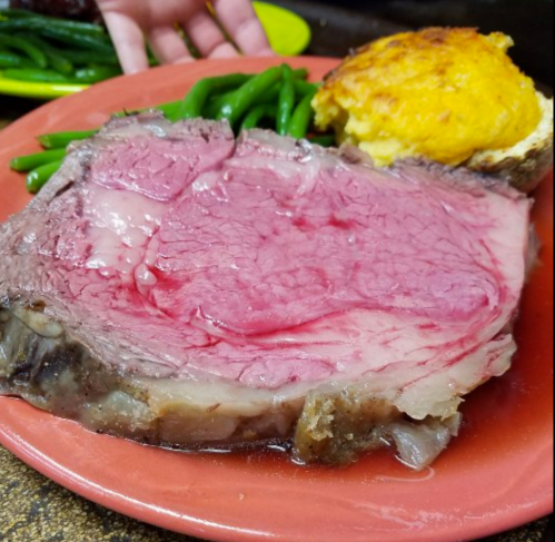 A plate featuring a large slice of rare prime rib, green beans, and a piece of cornbread.