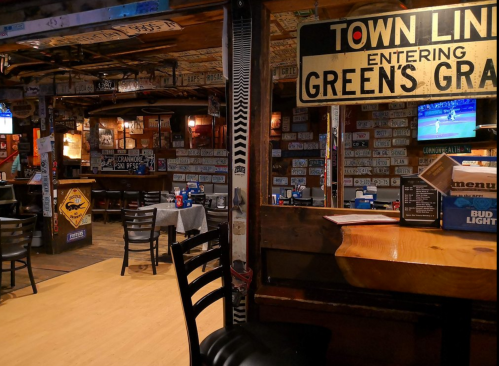 A cozy bar interior with wooden tables, chairs, and walls adorned with license plates and a TV showing a game.