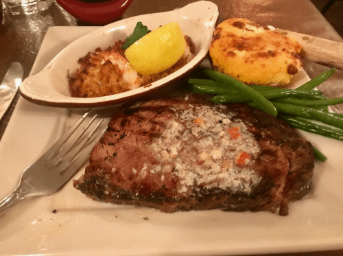 A plate featuring a grilled steak, lemon, green beans, a baked potato, and shrimp with a side of cornbread.