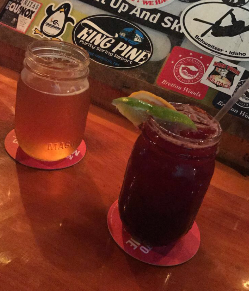 Two drinks on a wooden table: a light beer in a jar and a dark cocktail with a lime wedge. Stickers in the background.