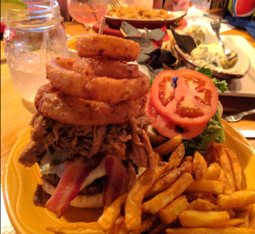 A hearty burger topped with pulled pork, bacon, onion rings, lettuce, and tomato, served with fries on a yellow plate.