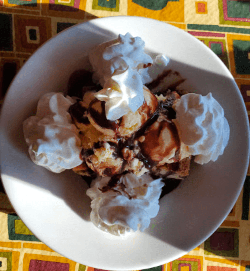 A bowl of ice cream topped with whipped cream and chocolate sauce, served on a colorful patterned tablecloth.