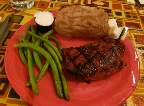 A red plate with a grilled steak, baked potato, green beans, and a small cup of sour cream.