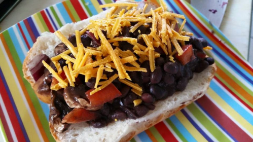 A colorful plate holds a sandwich topped with black beans, diced apples, and shredded cheese on a soft bread roll.