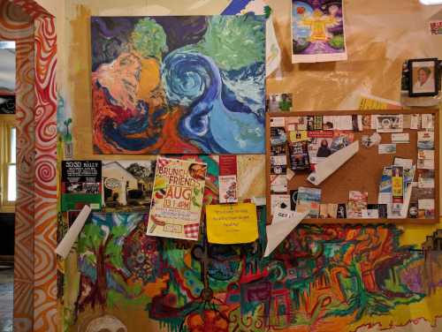 Colorful abstract painting on a wall, surrounded by flyers, a bulletin board, and vibrant mural art.