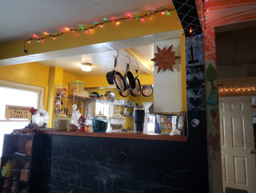 A cozy kitchen with colorful walls, hanging pots, and festive lights, featuring a chalkboard and various glass containers.