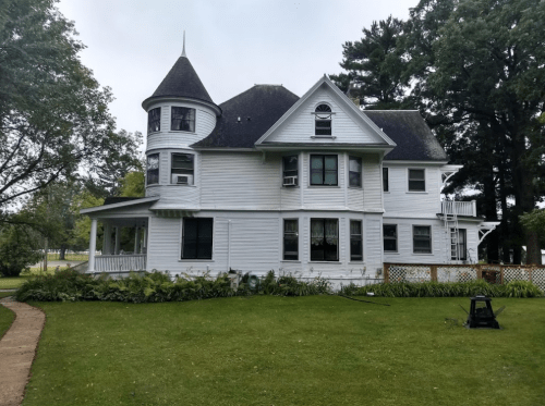 A large, white Victorian-style house with a turret, surrounded by greenery and a well-maintained lawn.