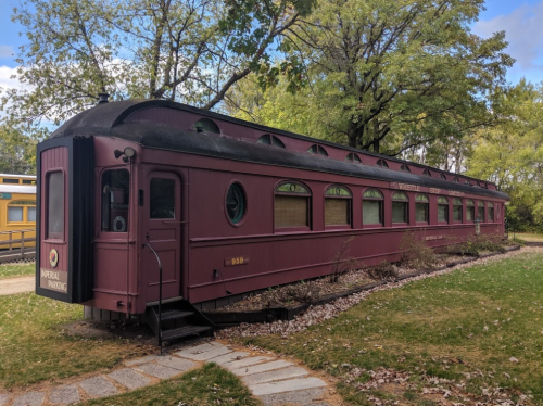 A vintage maroon train car converted into a building, surrounded by greenery and a stone pathway.