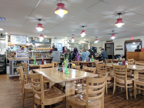 A cozy diner interior with wooden tables, red pendant lights, and customers at the counter and tables.
