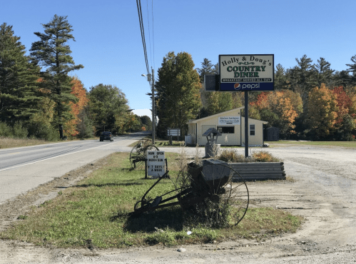 A roadside diner sign with autumn trees in the background, featuring a vintage farm implement in the foreground.