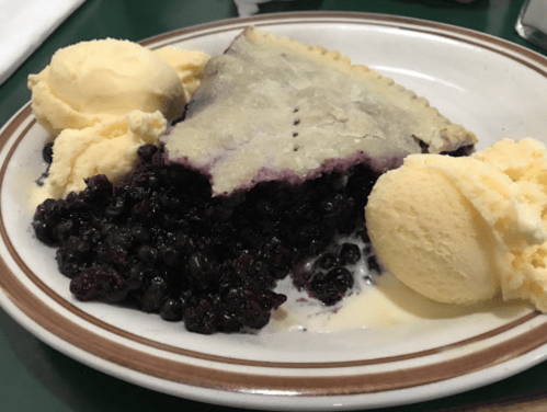A slice of blueberry pie with a flaky crust, served with two scoops of vanilla ice cream on a white plate.