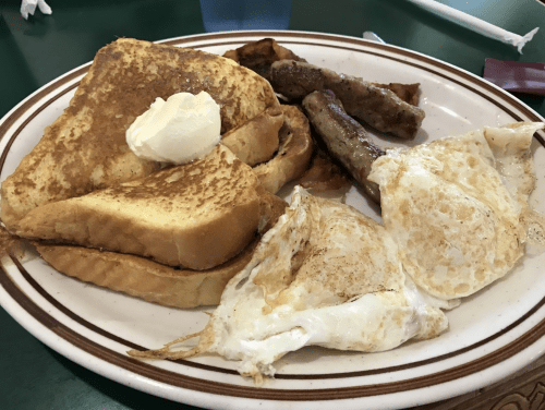 A plate of breakfast featuring French toast with butter, sausage links, and two fried eggs.