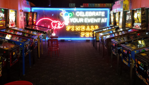 A vibrant arcade with rows of pinball machines and a neon sign that reads "Celebrate Your Event at Pinball Fest."