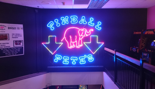 Neon sign for "Pinball Pete's" featuring an elephant and arrows pointing down, set against a dark wall.