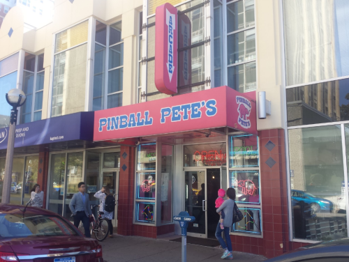 Exterior of Pinball Pete's arcade with bright signage, people walking by, and a bike parked outside.
