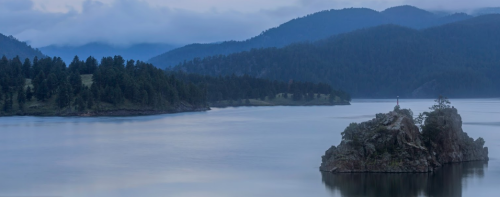 A serene lake surrounded by mountains, featuring a small rocky island with a lighthouse in the distance under a cloudy sky.