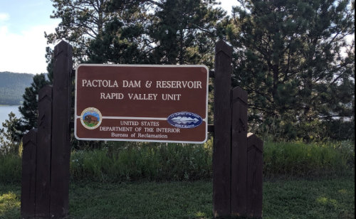 Sign for Pactola Dam & Reservoir, Rapid Valley Unit, surrounded by trees and water in the background.