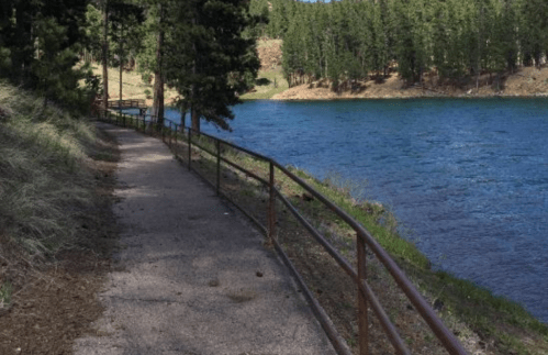 A scenic path along a lake, bordered by a railing and surrounded by trees and greenery.