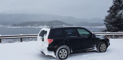 A black SUV parked on a snowy road with a lake and mountains in the background under a cloudy sky.