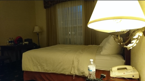 A hotel room with a bed, lamp, water bottle, and a desk by a window with sheer curtains.