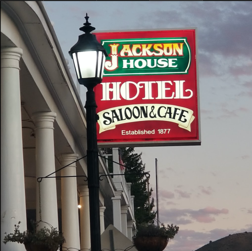 Sign for Jackson House Hotel, Saloon & Cafe, established 1877, with a lamppost in front and a colorful sunset backdrop.
