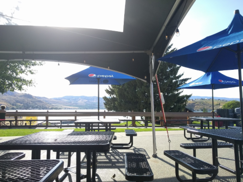Outdoor seating area with Pepsi umbrellas overlooking a lake and mountains under a bright sky.
