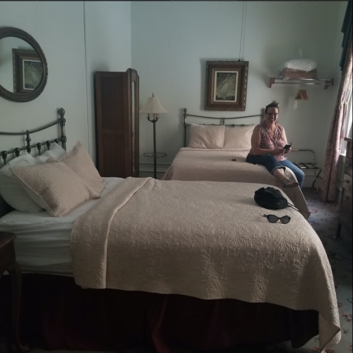 A cozy hotel room with two beds, a woman sitting by the window, and vintage decor.