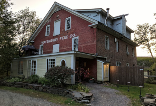 Historic building with red brick and yellow siding, labeled "Waterbury Feed Co," surrounded by trees and a gravel path.