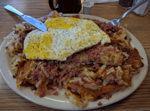 A plate of hash browns topped with corned beef and a fried egg, served with a knife and fork.