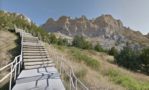 A wooden staircase leads up a grassy hill towards rocky cliffs under a clear blue sky.