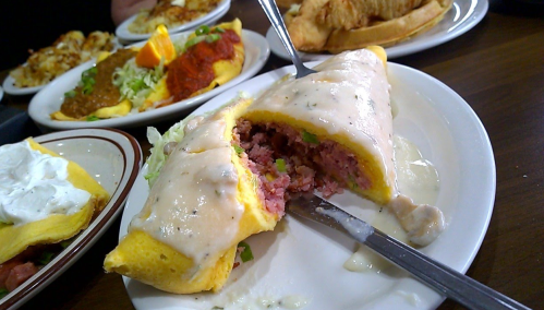 A plate with a cut open omelet filled with meat, topped with creamy sauce, alongside other dishes in the background.