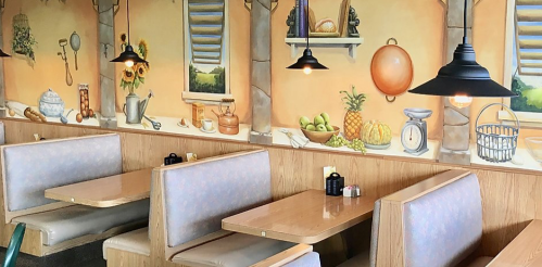 Cozy restaurant interior with wooden booths and a mural of kitchen items and fruits on the wall. Warm lighting.