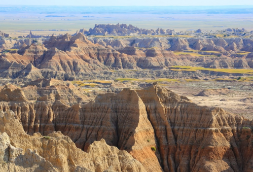 A panoramic view of rugged, layered rock formations and valleys in a vast, arid landscape under a clear blue sky.