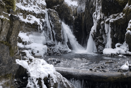 A snowy landscape featuring a waterfall cascading into a frozen pool, surrounded by icy rocks and frosted trees.
