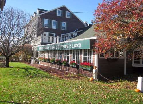 A charming restaurant with a green sign, surrounded by colorful flowers and autumn foliage, set against a clear blue sky.