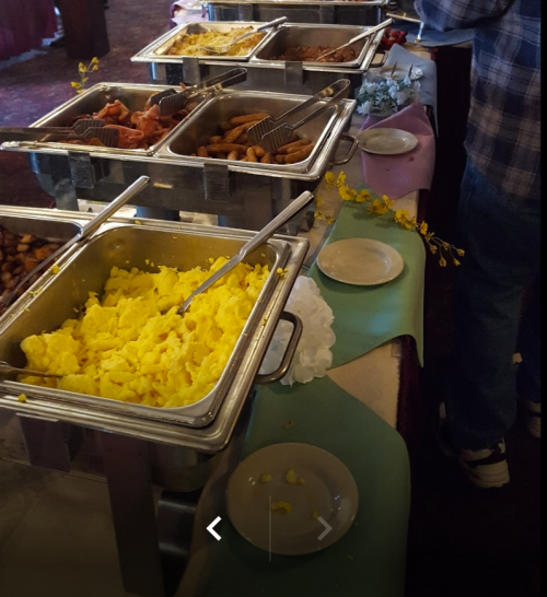 A buffet table with trays of scrambled eggs, meats, and sides, set up for a meal in a decorated venue.