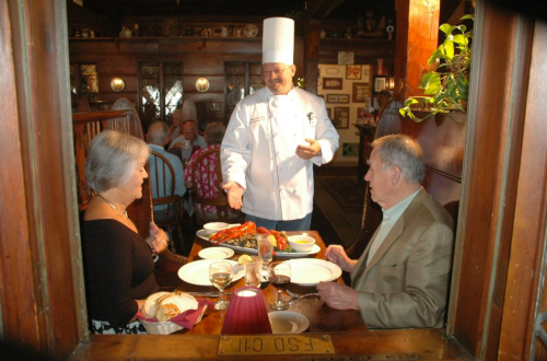 A chef presents a seafood dish to a couple at a cozy restaurant, with other diners in the background.